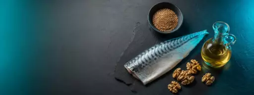 Whole mackerel alongside walnuts, a small bowl of lentils, and a carafe of olive oil on a dark slate surface