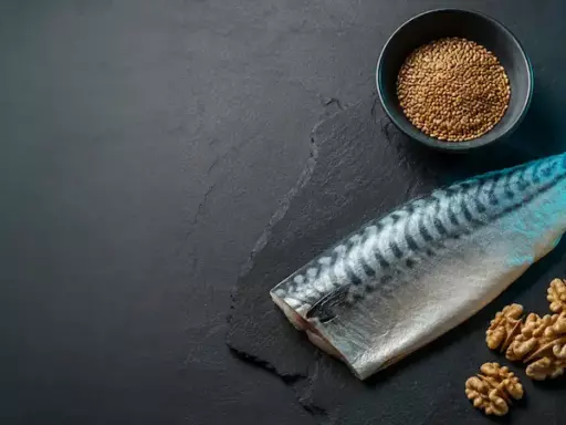 Whole mackerel alongside walnuts, a small bowl of lentils, and a carafe of olive oil on a dark slate surface