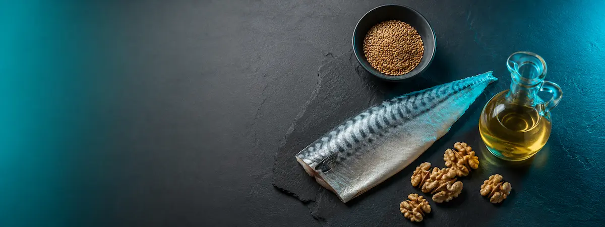 Whole mackerel alongside walnuts, a small bowl of lentils, and a carafe of olive oil on a dark slate surface