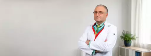 Male doctor in a white lab coat with arms crossed, standing in a bright room with a plant on a shelf in the background.