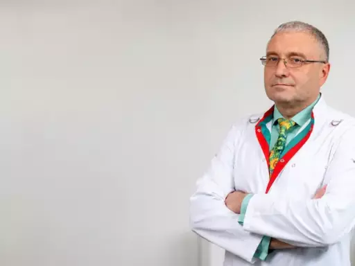 Male doctor in a white lab coat with arms crossed, standing in a bright room with a plant on a shelf in the background.