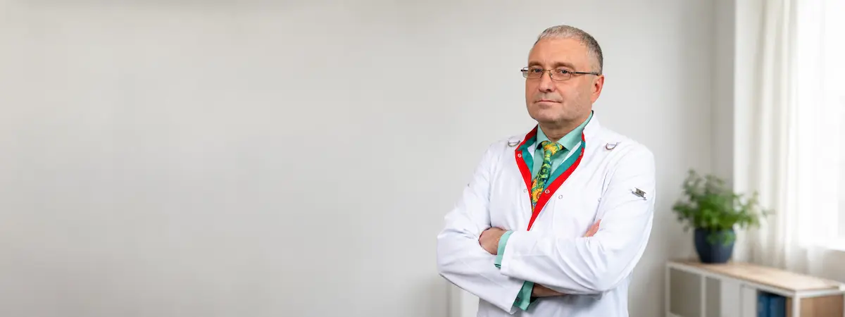 Male doctor in a white lab coat with arms crossed, standing in a bright room with a plant on a shelf in the background.