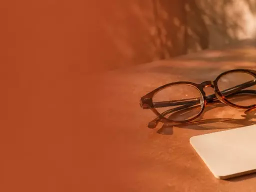 Brown ceramic cup, a pair of round glasses, and a blank card on a warm-toned tabletop in soft sunlight.