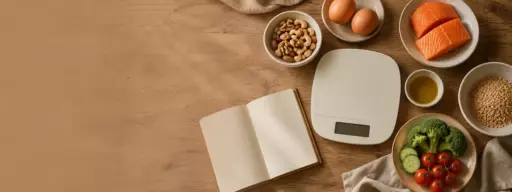 Open recipe notebook on a wooden kitchen table beside a digital scale, with bowls of salmon, eggs, nuts, seeds, olive oil, and a veggie bowl.